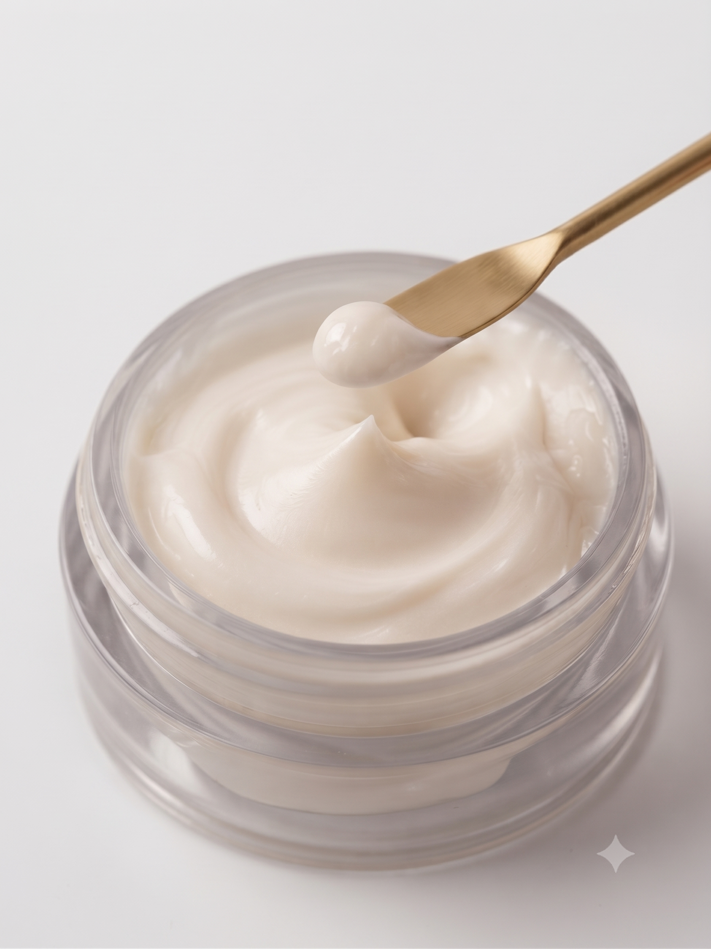 A close-up of a clear cosmetic jar filled with thick, creamy off-white cream, with a gold spatula lifting a dollop of the product against a clean white background.