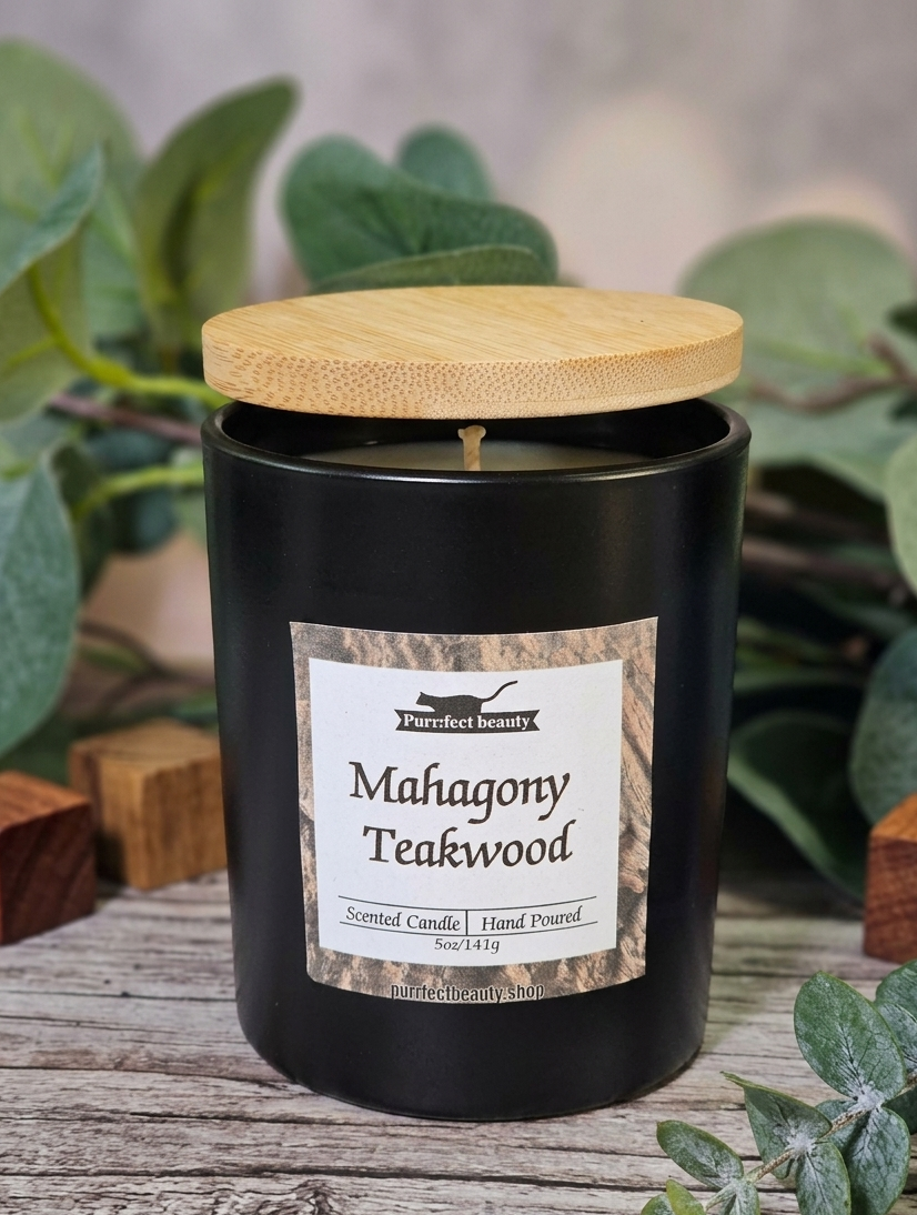 Black candle with wooden lid labeled 'Mahogany Teakwood' on a wooden surface with greenery.