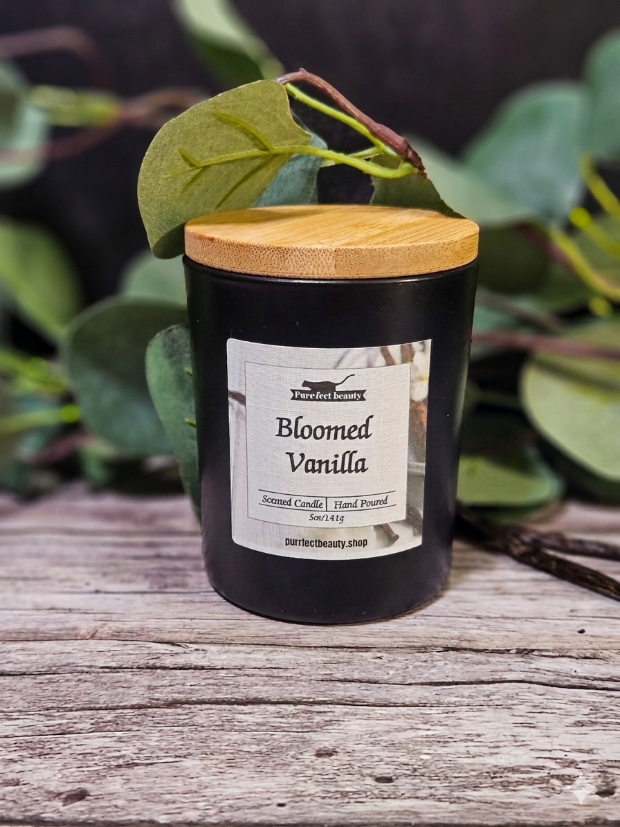 Black candle labeled 'Bloomed Vanilla' with a wooden lid on a wooden surface with green foliage in the background.