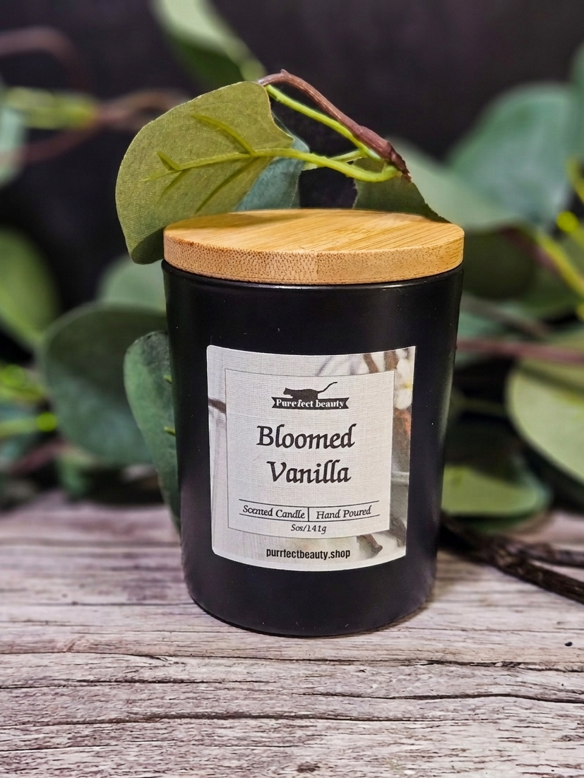 Black candle labeled 'Bloomed Vanilla' with a wooden lid on a wooden surface with green foliage in the background.
