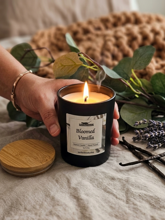 Hand holding a lit candle labeled 'Bloomed Vanilla' with green leaves and lavender in the background.