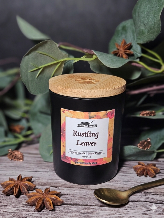 Black candle with a wooden lid labeled 'Rustling Leaves' surrounded by green leaves and star anise on a dark background.