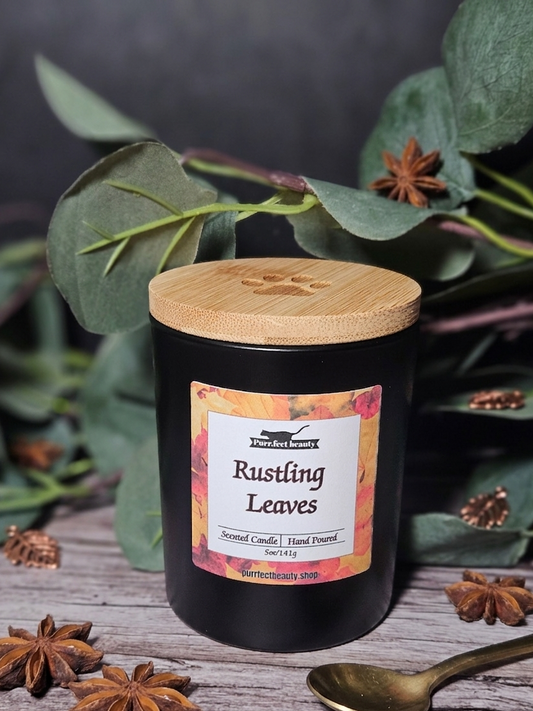Black candle with a wooden lid labeled 'Rustling Leaves' surrounded by green leaves and star anise on a dark background.