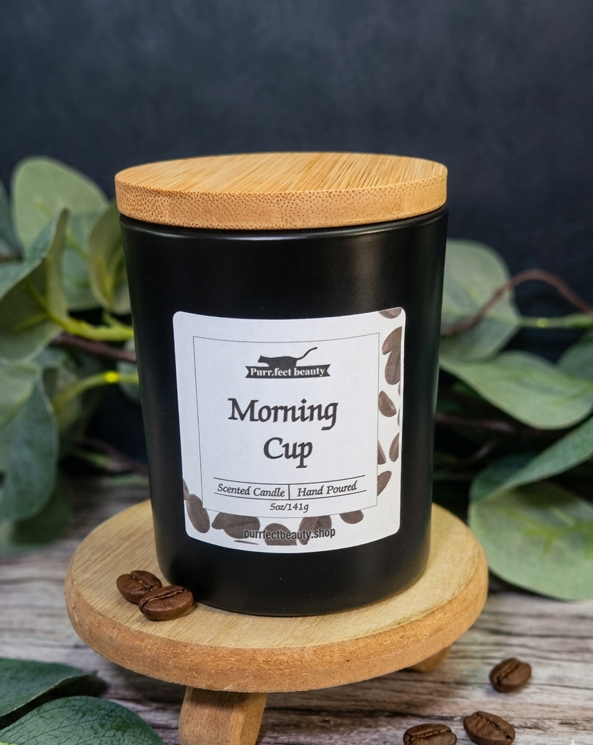 Black candle labeled 'Morning Cup' with a wooden lid on a wooden stand, surrounded by coffee beans and leaves.