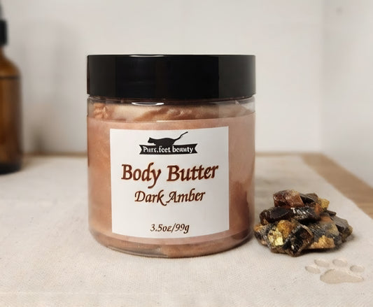 A jar of Pure Feet Beauty Body Butter in the Dark Amber scent, featuring a black lid and a white label, set on a light fabric surface next to a small pile of dark amber crystals.