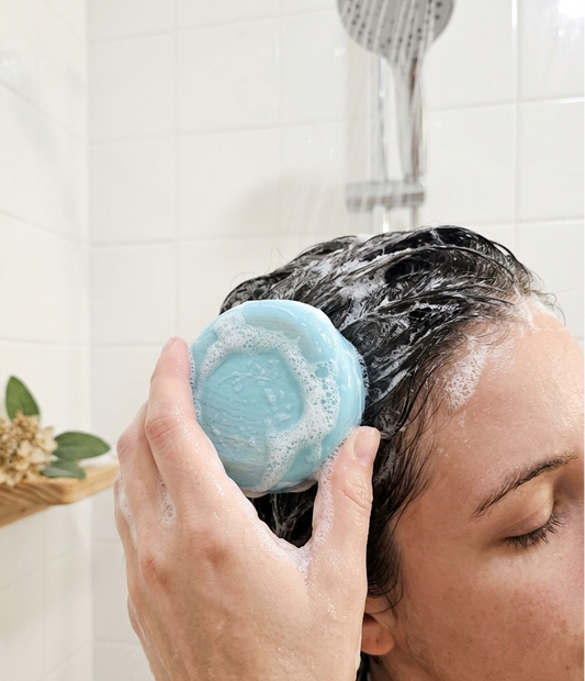 Person using a blue circular shampoo bar on their hair in a bathroom setting.