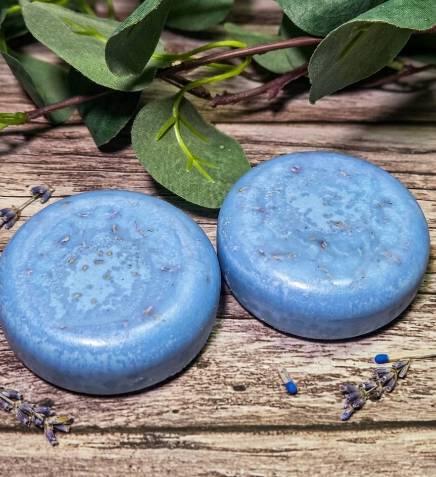 Two blue shampoo bars on a wooden surface with green leaves and lavender flowers.