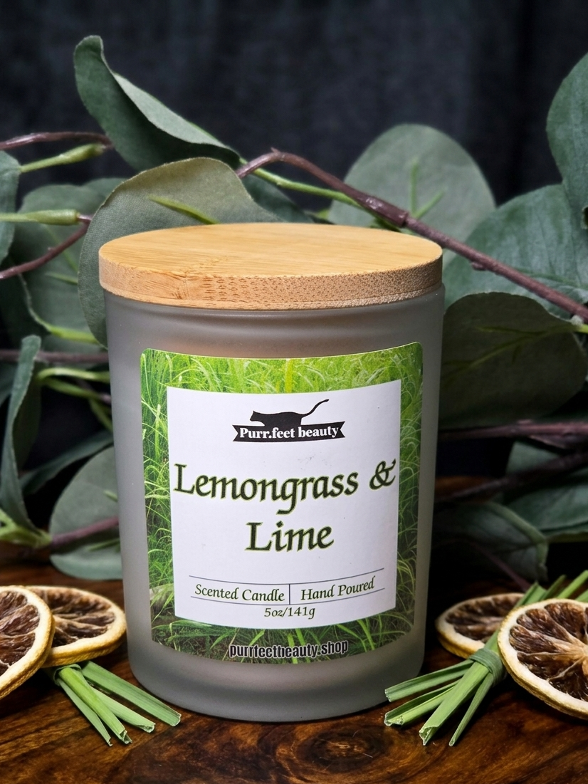 Candle labeled 'Lemongrass & Lime' with a wooden lid, surrounded by green leaves and dried oranges on a dark background.