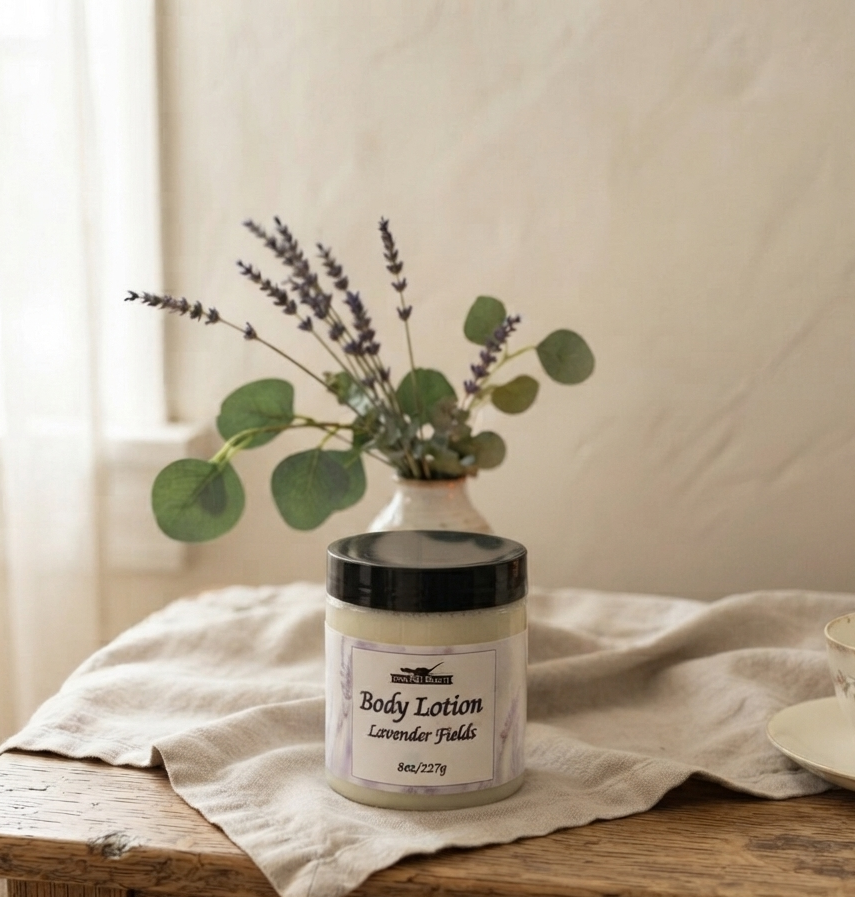 A jar of body lotion labeled 'Body Lotion Lavender Fields' sits on a rustic wooden table draped with a cream linen cloth, styled with a small vase of lavender and eucalyptus leaves, with a floral teacup in the background.