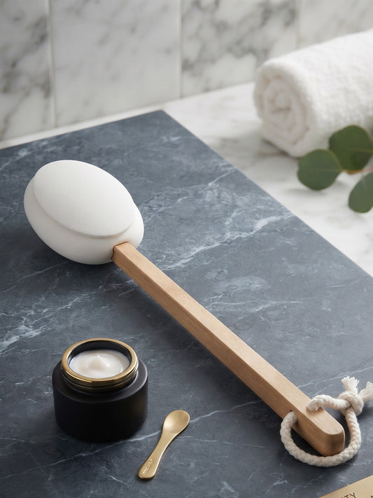 A spa-quality back lotion applicator featuring a white silicone backer, a natural wood handle, a white rope loop, a small gold metal spoon, and a black jar of lotion on a dark gray marble tray.