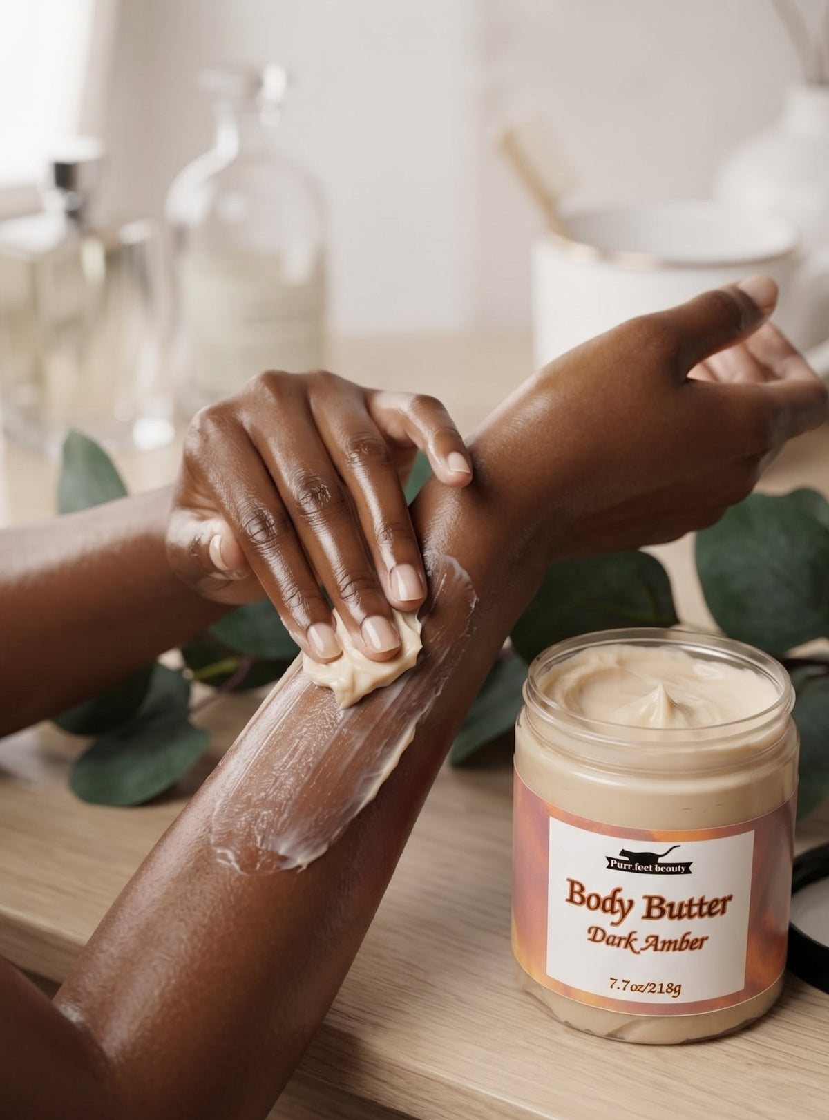 A person applies a white body butter cream to their forearm next to an open jar of 'Body Butter Dark Amber' by The Body Shop, set on a wooden table with soft-focus background elements.