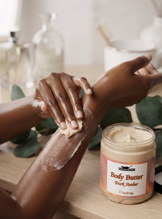 A person applies a white body butter cream to their forearm next to an open jar of 'Body Butter Dark Amber' by The Body Shop, set on a wooden table with soft-focus background elements.