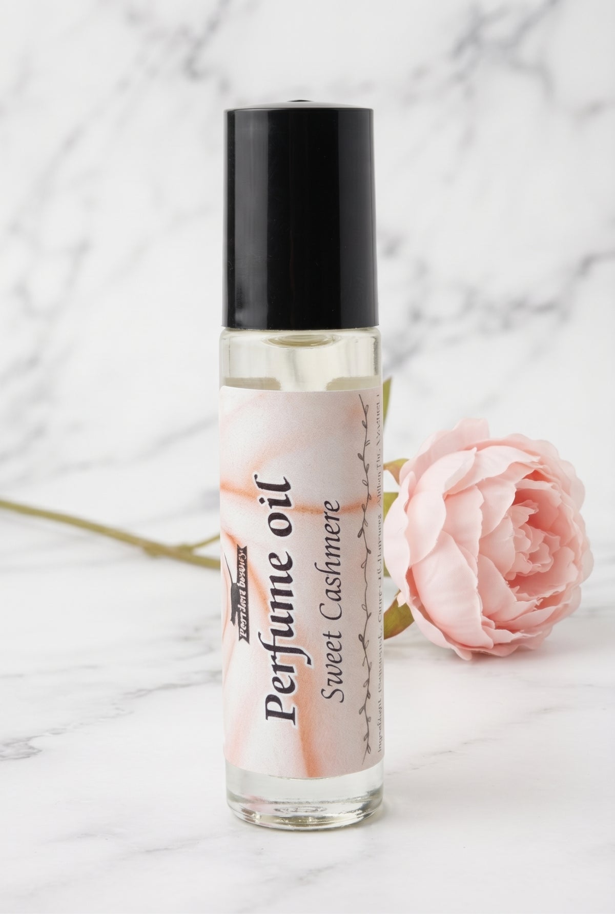 A clear glass roll-on perfume oil bottle with a black cap and a white label reading 'Perfume oil Sweet Cashmere', set on a white marble surface next to a pink rose.
