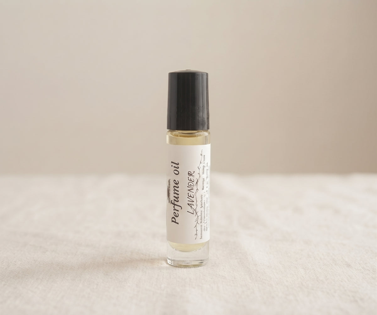 A small clear glass perfume oil roller bottle with a black cap and a white label reading 'Perfume oil Lavender', standing on a light fabric surface against a soft beige background.