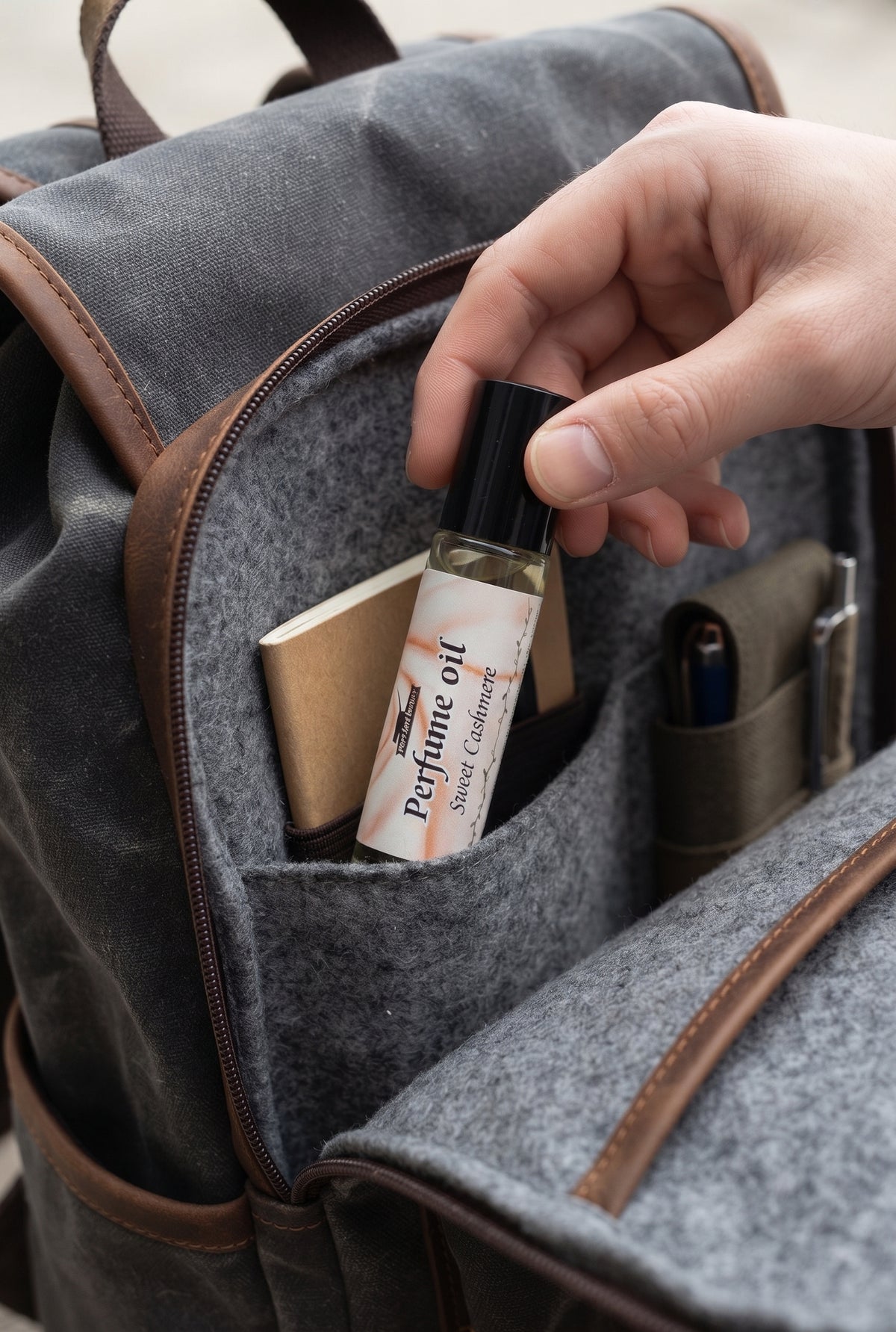 A close-up of a hand placing a small perfume oil bottle into a gray felt backpack pocket with brown leather trim.
