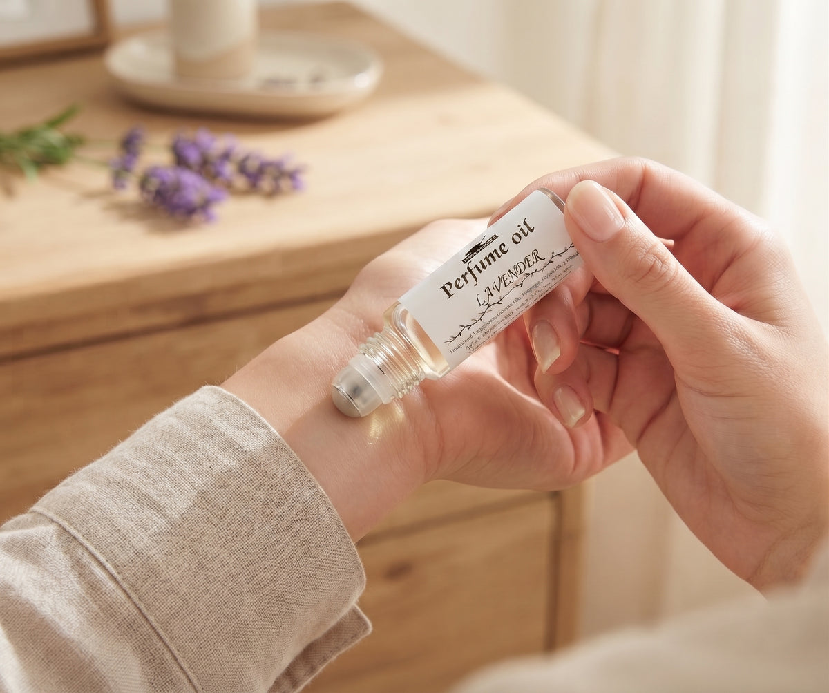 A close-up of a person's hand holding a small glass bottle of lavender perfume oil with a silver rollerball, set against a warm, softly blurred background of a wooden table and curtains.