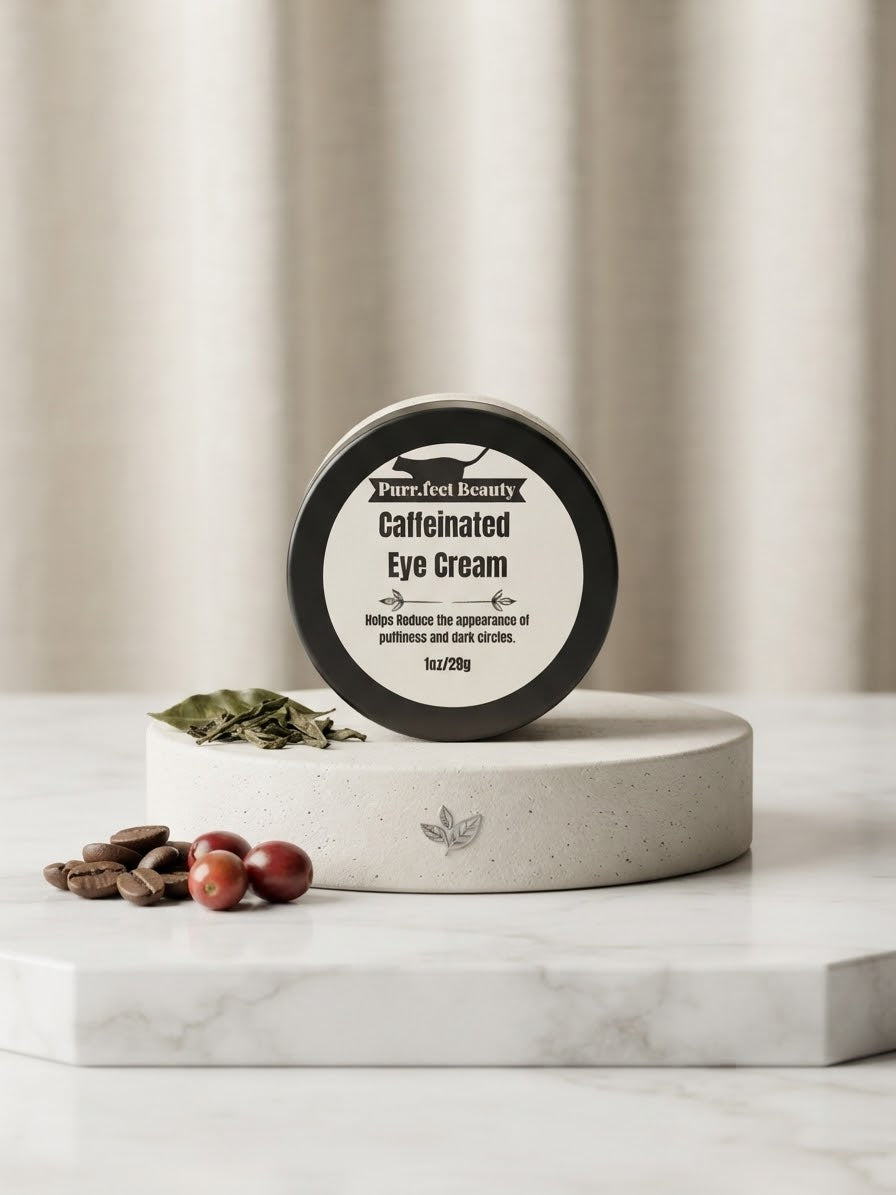 A round black-and-white cosmetic jar labeled 'Purrfect Beauty Caffeinated Eye Cream' sits on a white pedestal with coffee beans and dried leaves nearby on a marble surface.