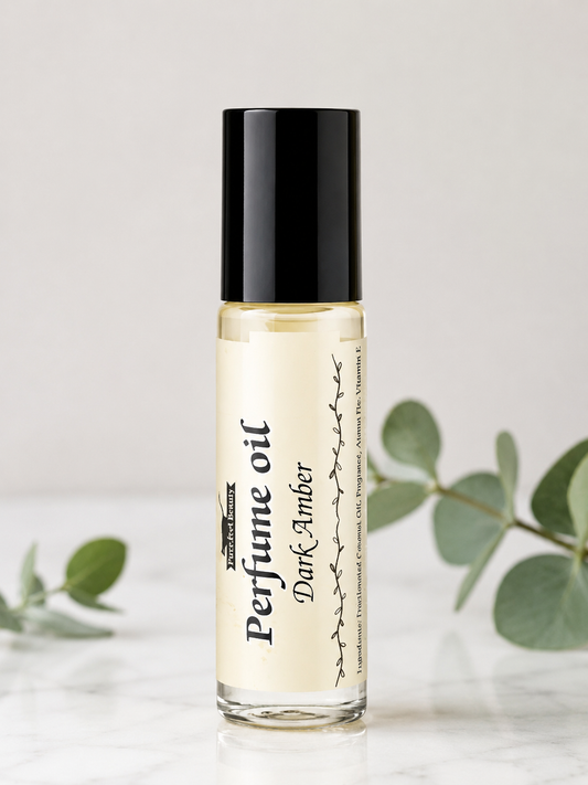A clear glass bottle of perfume oil with a black cap and a cream label reading 'Perfume oil Dark Amber', set on a light marble surface with soft green leaves in the background.