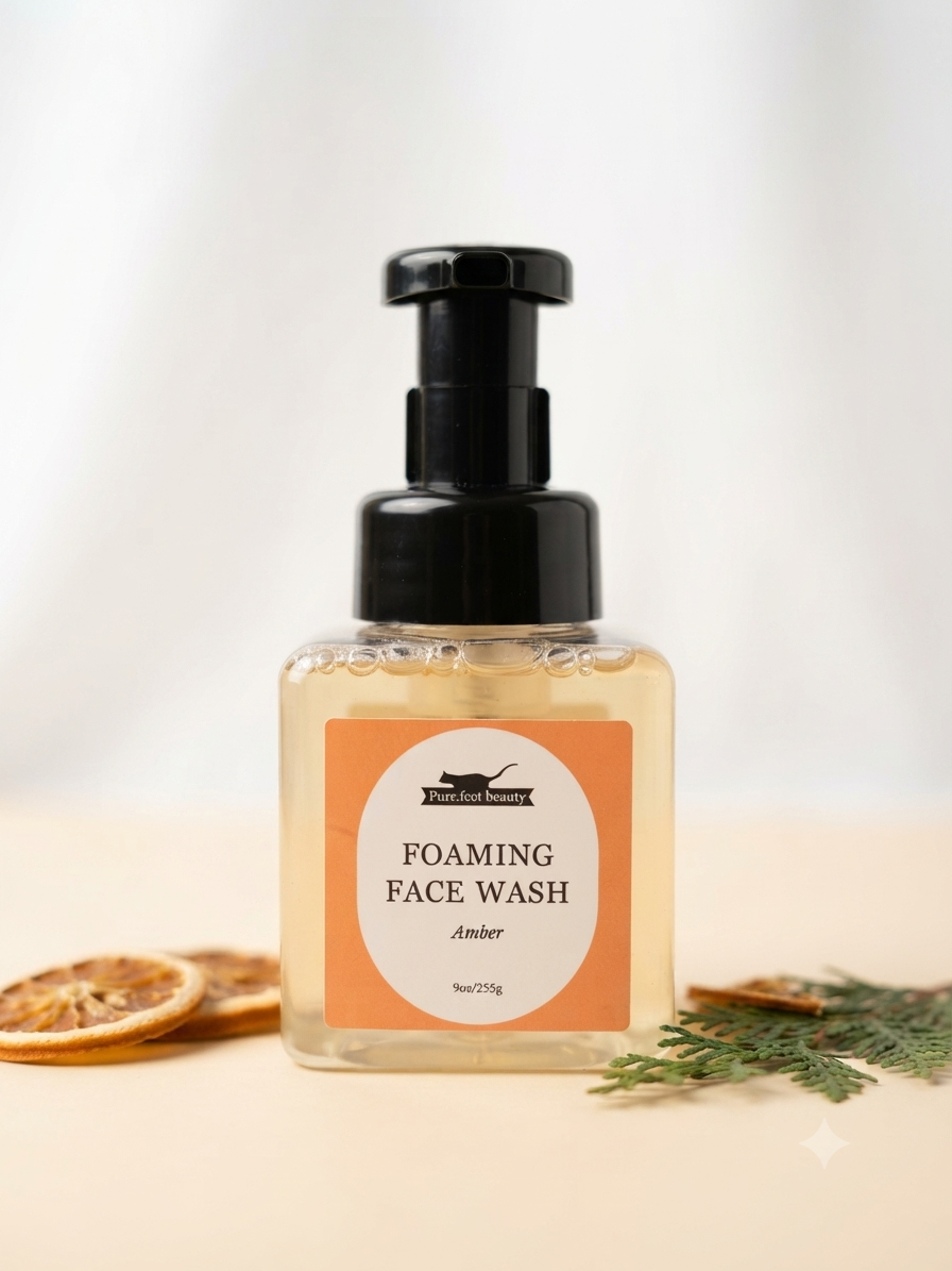 A bottle of Purr.fect beauty amber-colored foaming face wash with a black pump dispenser and an orange label, styled on a light beige surface with dried orange slices and greenery.
