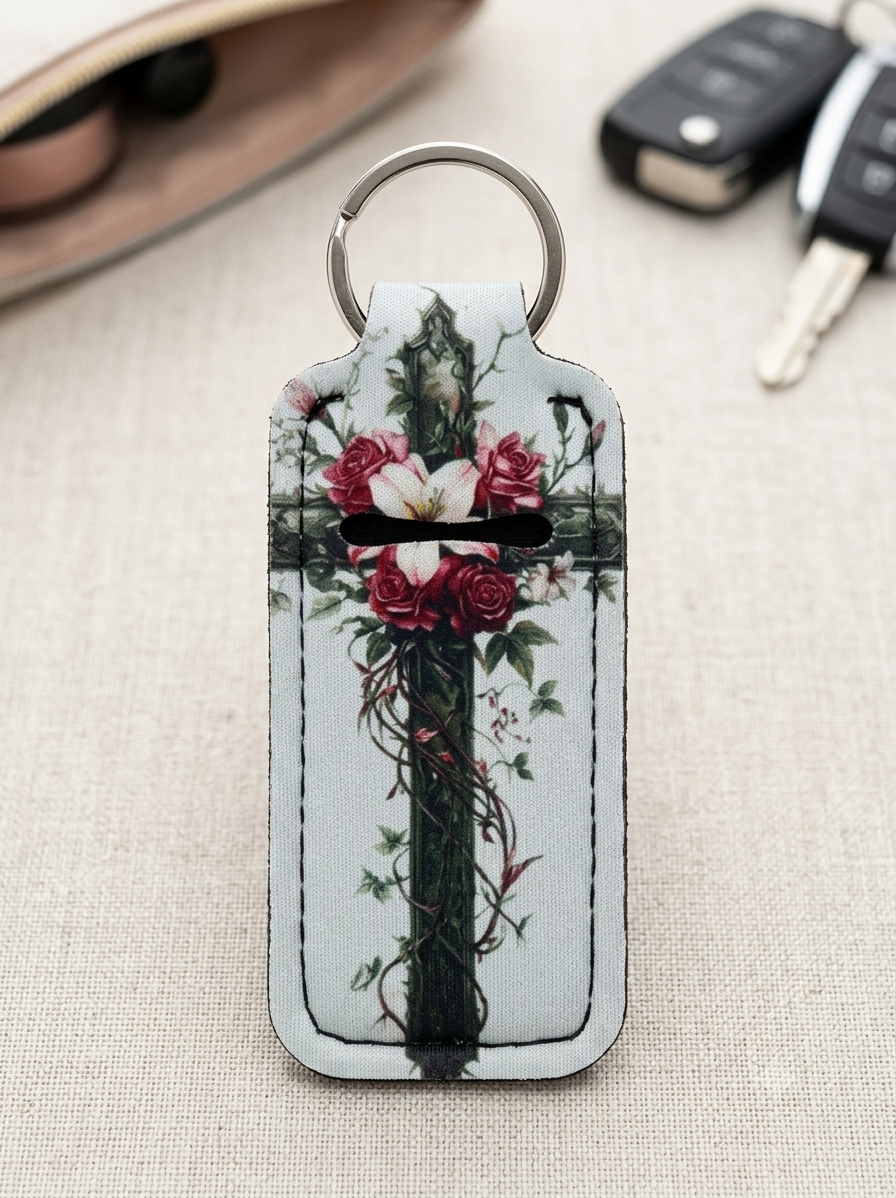 A rectangular leather keychain wallet with a silver split ring, featuring a printed design of a dark green cross wrapped in red roses and green vines on a light background. The keychain is displayed on a beige textured fabric surface with blurred sunglasses and car keys in the background.
