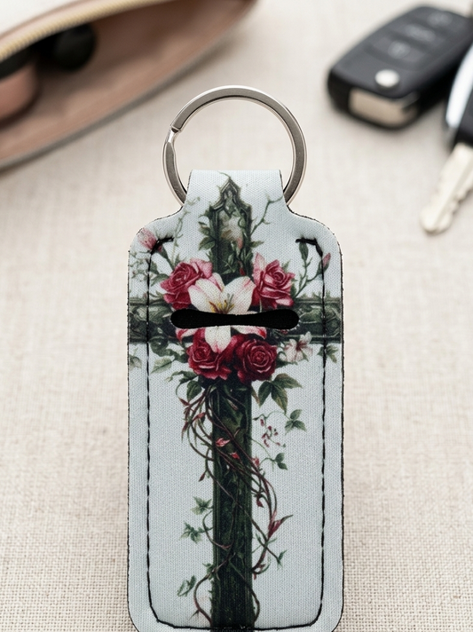 A rectangular leather keychain wallet with a silver split ring, featuring a printed design of a dark green cross wrapped in red roses and green vines on a light background. The keychain is displayed on a beige textured fabric surface with blurred sunglasses and car keys in the background.