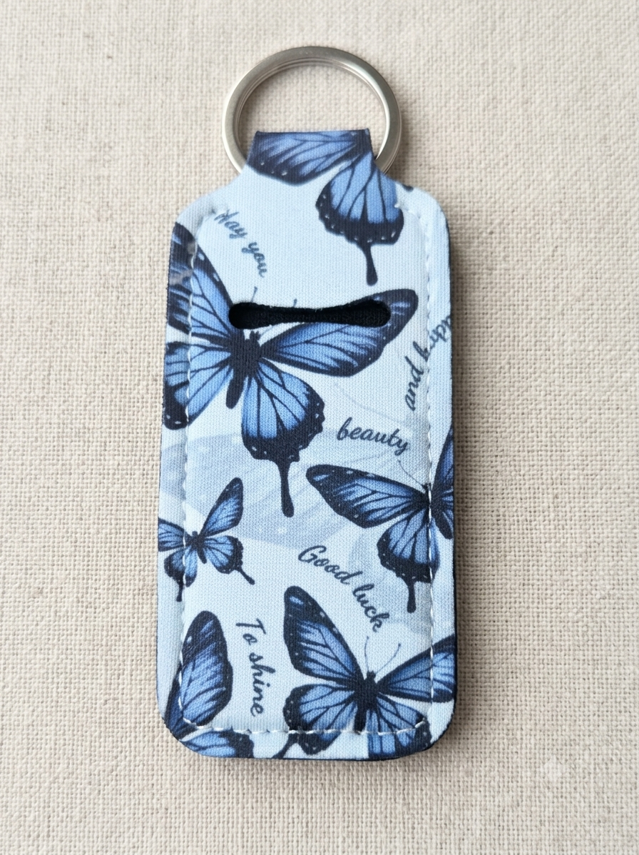 A fabric keychain wallet with a butterfly print in shades of blue and white, featuring a silver metal key ring and a coin slot, set against a neutral beige textured background.
