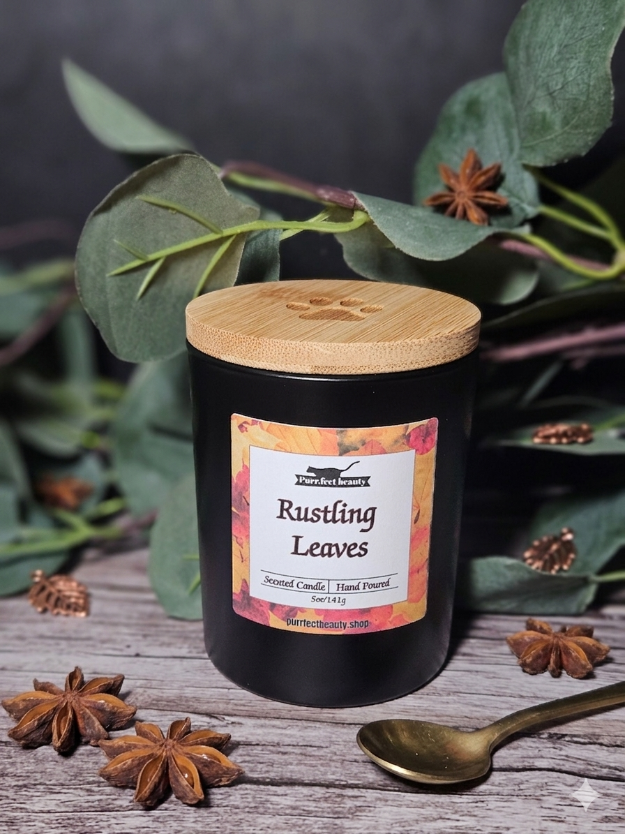 Black candle with a wooden lid labeled 'Rustling Leaves' surrounded by green leaves and star anise on a dark background.