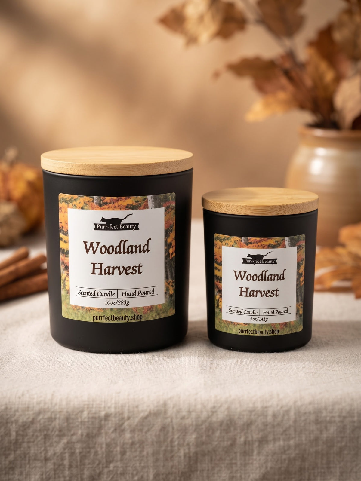 Two black scented candles with light wood lids labeled 'Woodland Harvest' by Perfectly Picky Shop, displayed on a linen surface with autumn leaves and a vase in the background.